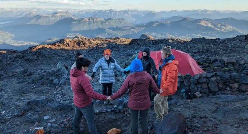 A group of people stand in a circle holding hands on top of a rocky overlook. Near them, there is a tent, and behind them is a vast mountainous landscape. 